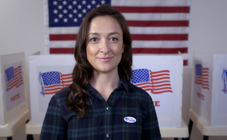 featured image showing American woman in plaid shirt wearing "I Voted" sticker and looking to camera, standing proud in front of polling booths with US flag