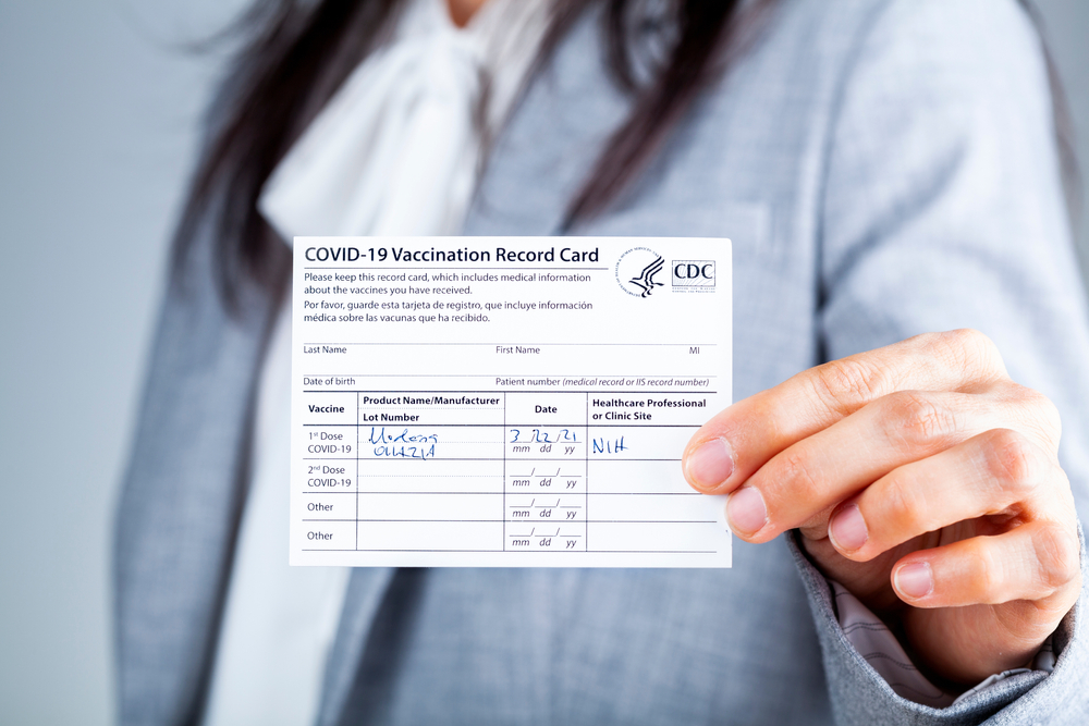 Featured image showing a A caucasian businesswoman is showing her CDC issued COVID vaccination record card as a proof of immunization. It has the date and lot number on it.
